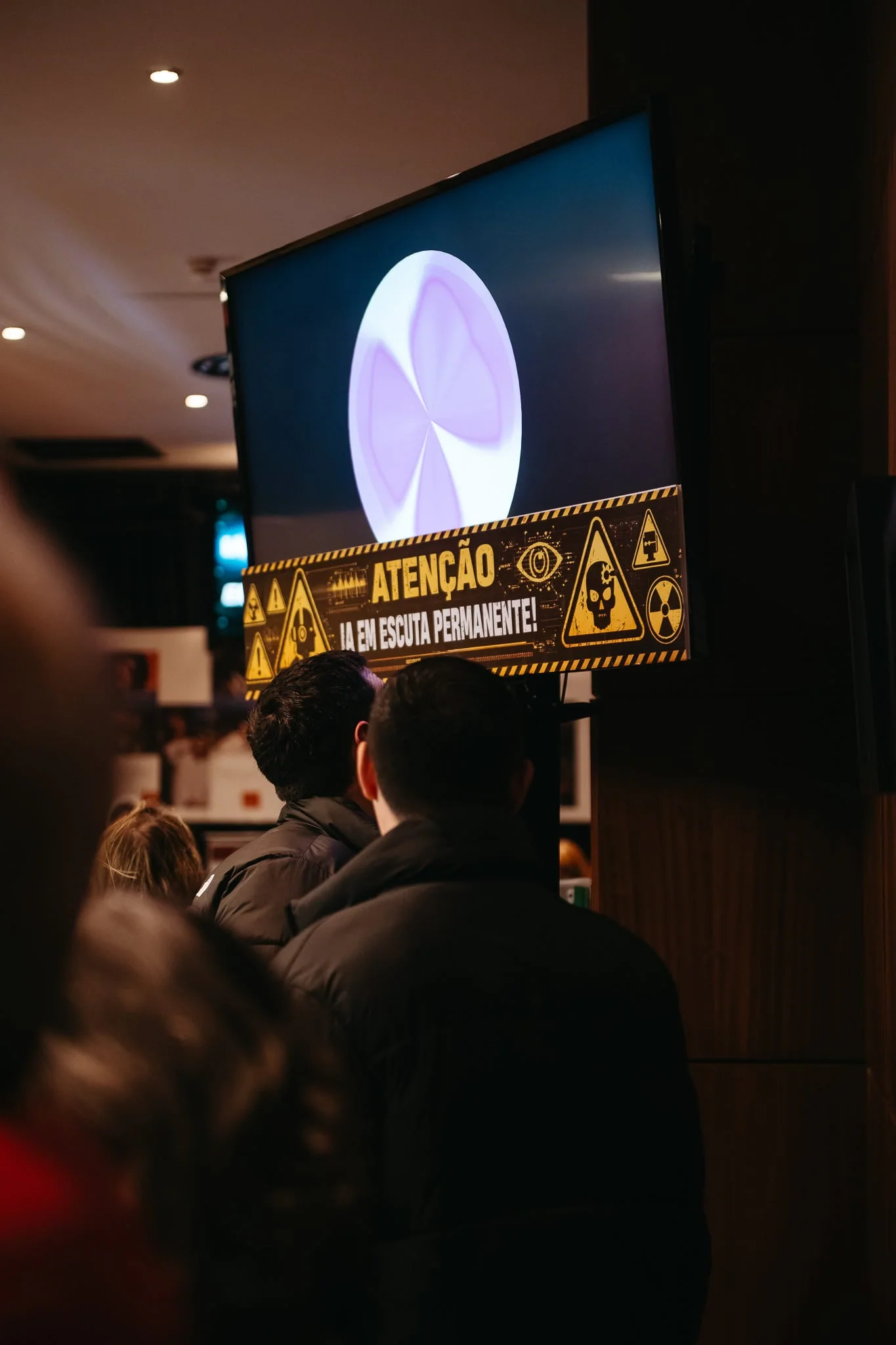 Audience members facing a screen that displays the AI character’s orb and a Portuguese listening notice.