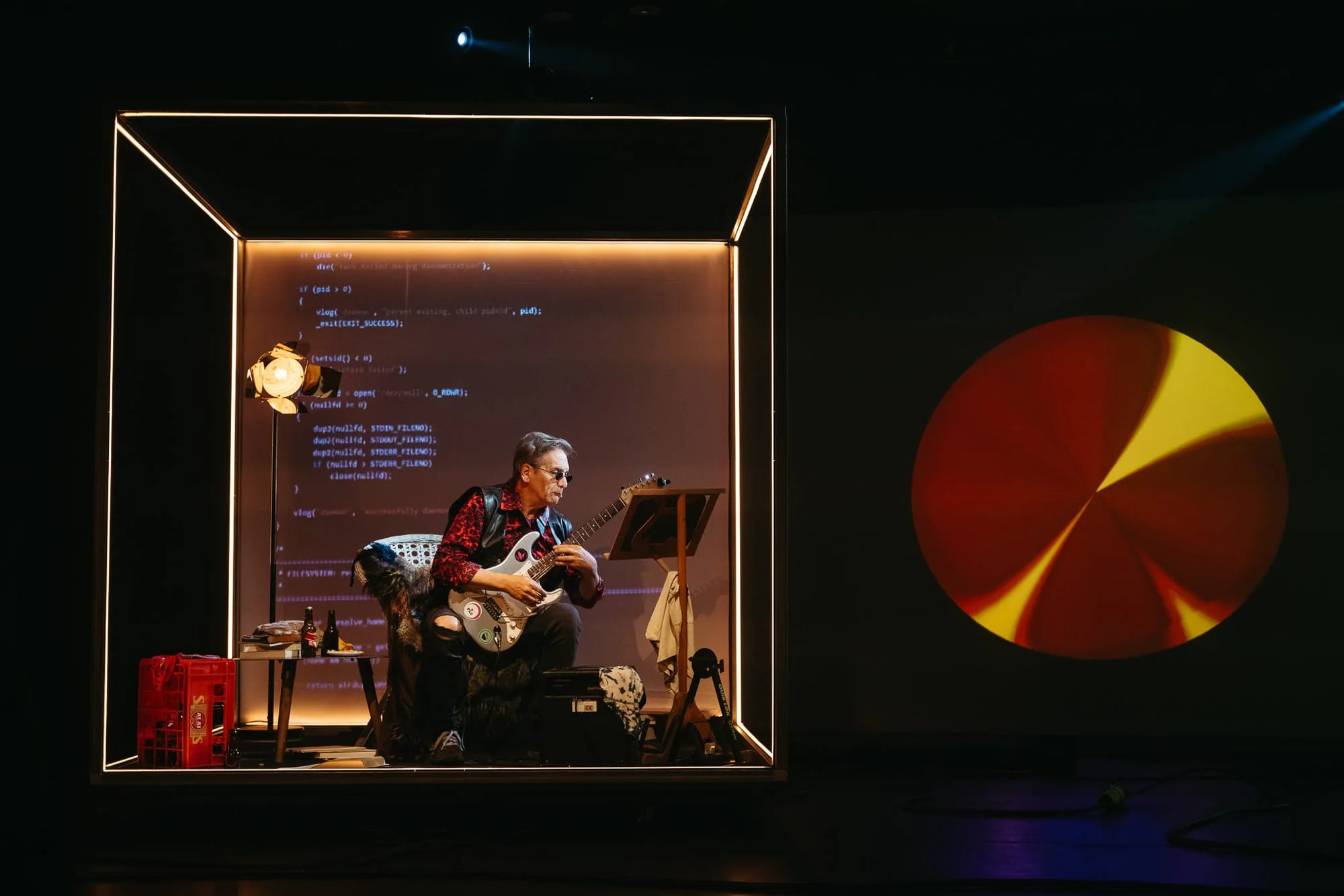 Performer playing guitar inside a lit stage installation, with a projected graphic beside him.