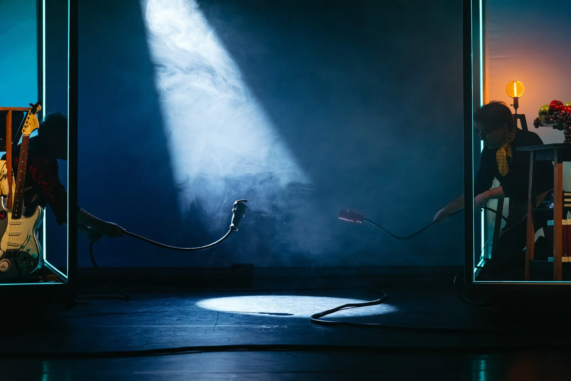 Dark theatre stage with a musician and a performer silhouetted around a central spotlight.